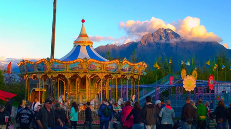 Fair goers surround a farris wheel and small roller coaster as the sun sets with Pioneer Peak in the background.